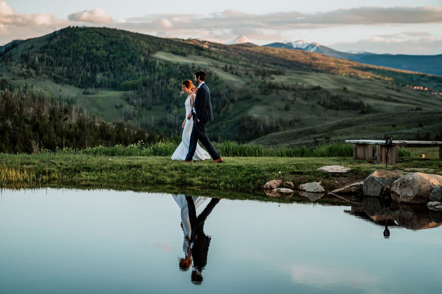 A scenic mountain wedding photo with couple's reflection in still water during sunset.