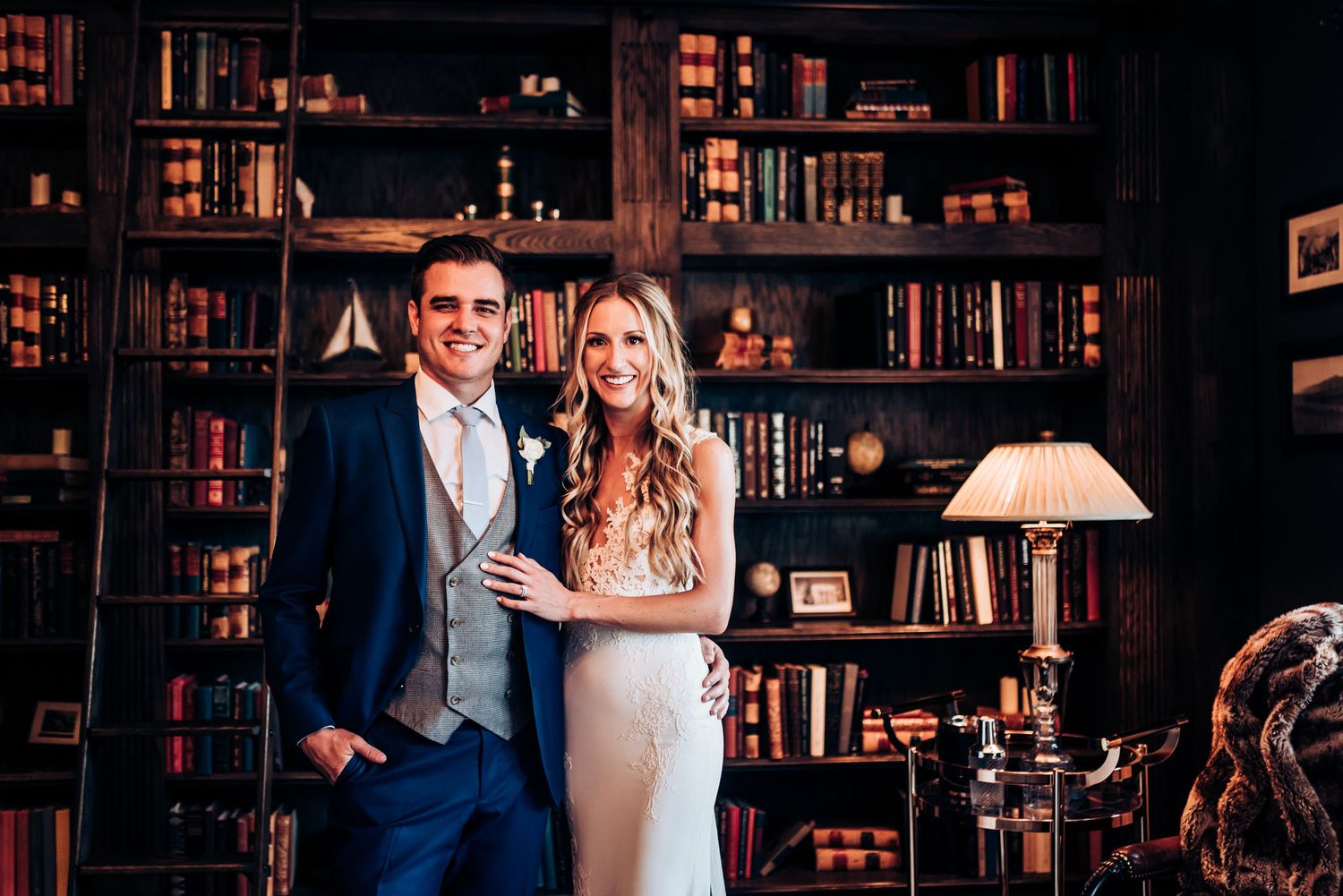 A wedding couple poses in formal attire against a backdrop of floor-to-ceiling bookshelves in a library.