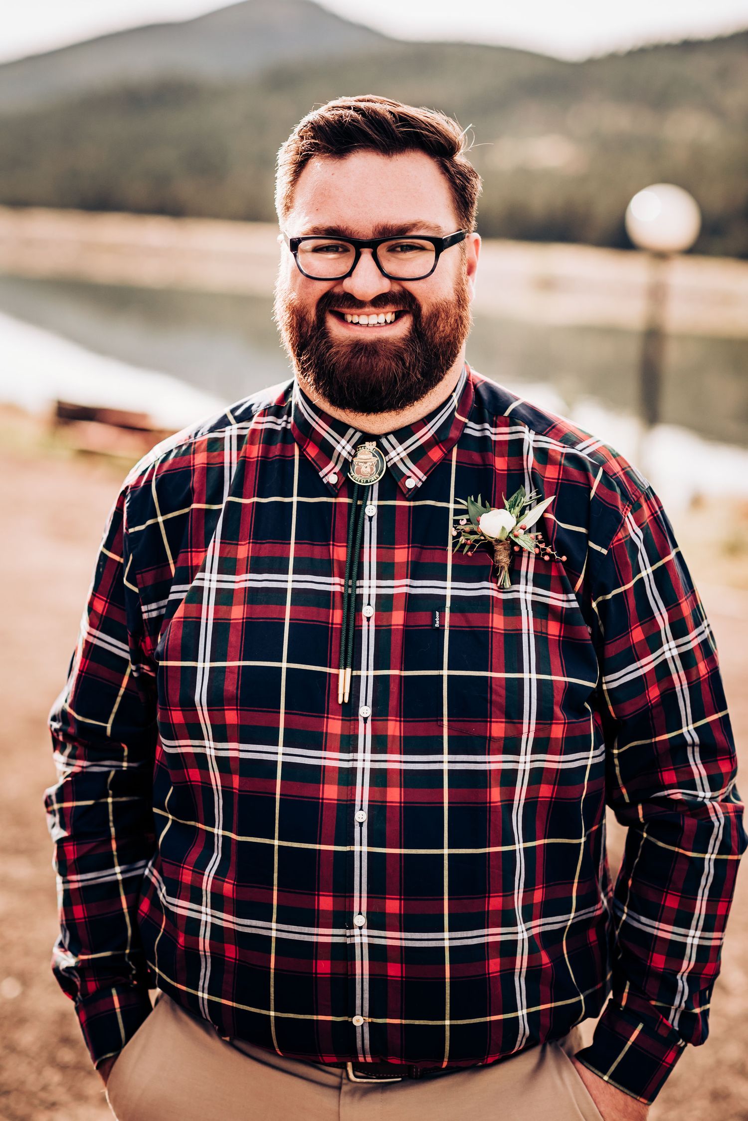 Person in red plaid button-down shirt with boutonniere smiles warmly against mountain backdrop.