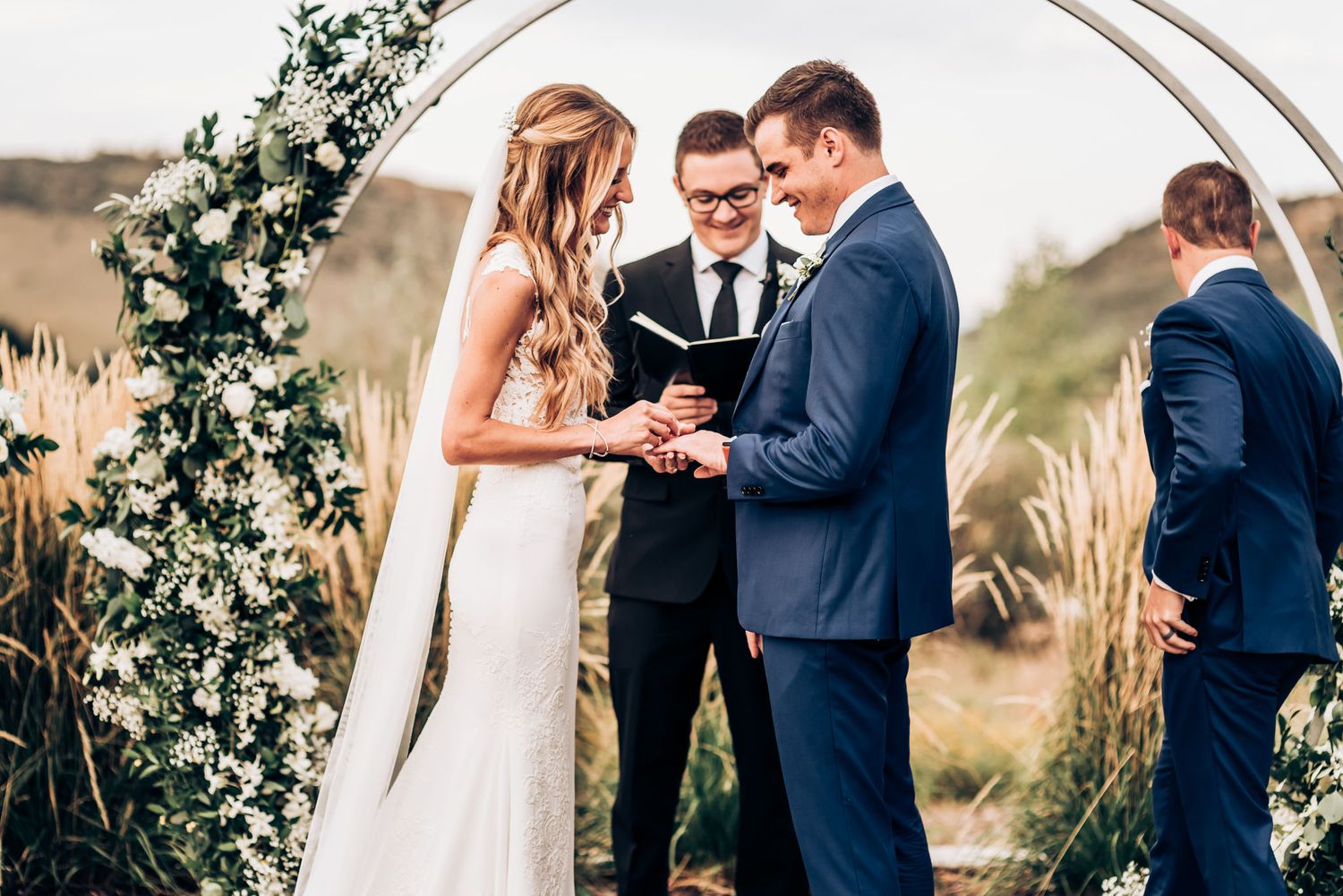 Exchange of rings at outdoor wedding beneath circular arch adorned with white flowers.