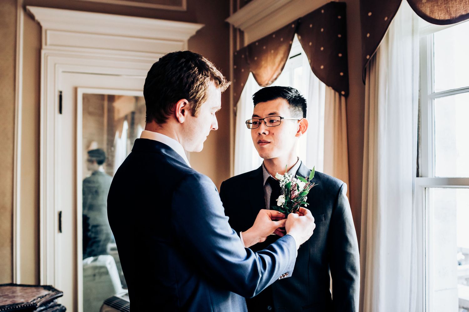 Two people in formal attire adjusting a boutonniere near a window.