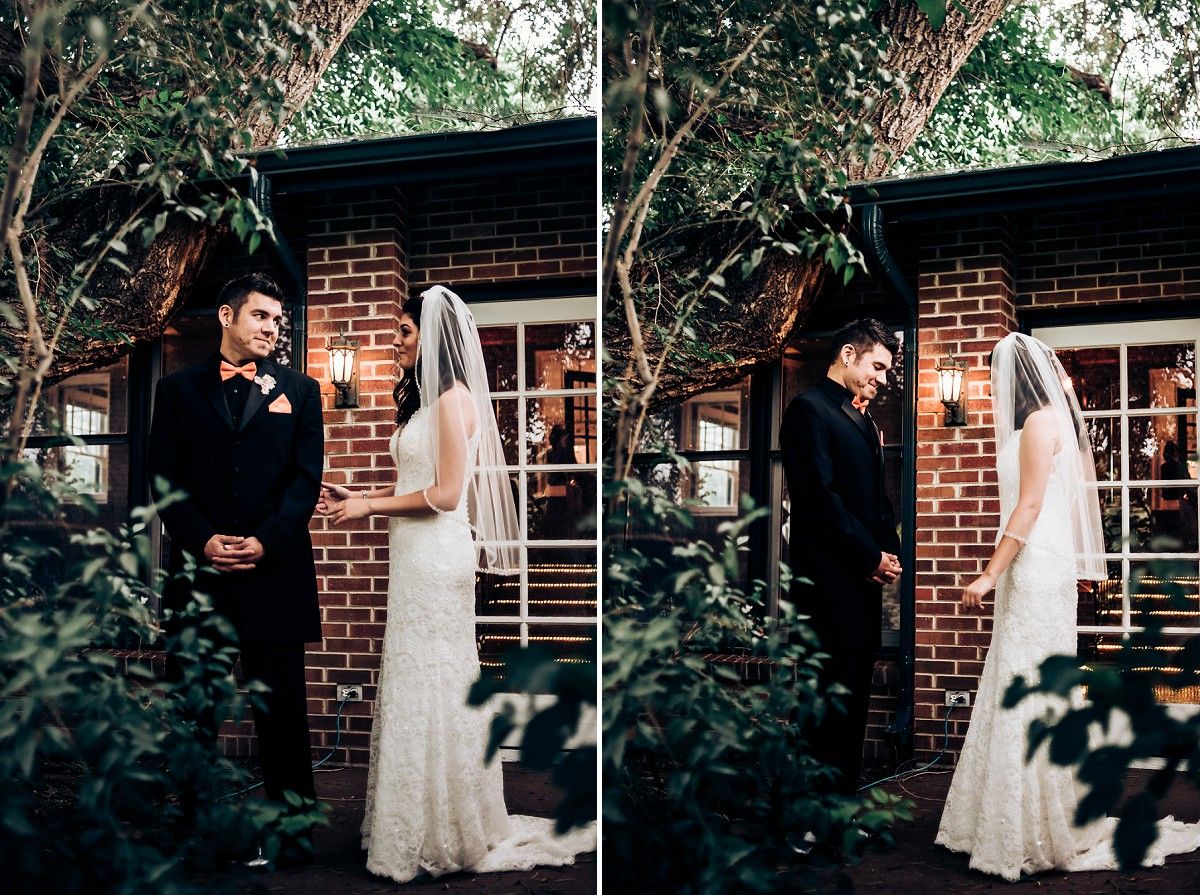 A romantic first look between a bride in white lace and groom in black suit against a brick building with greenery.