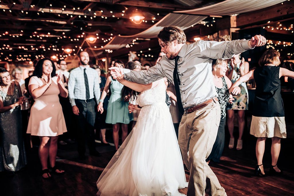 Dancing guests celebrate at a wedding reception beneath glowing string lights in a rustic venue.