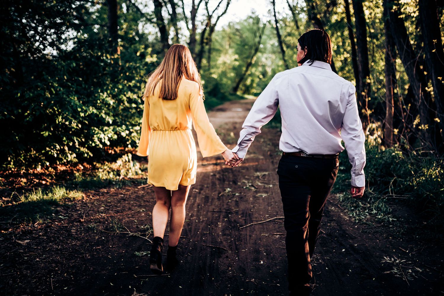 A couple walks hand in hand down a forest path during golden hour.