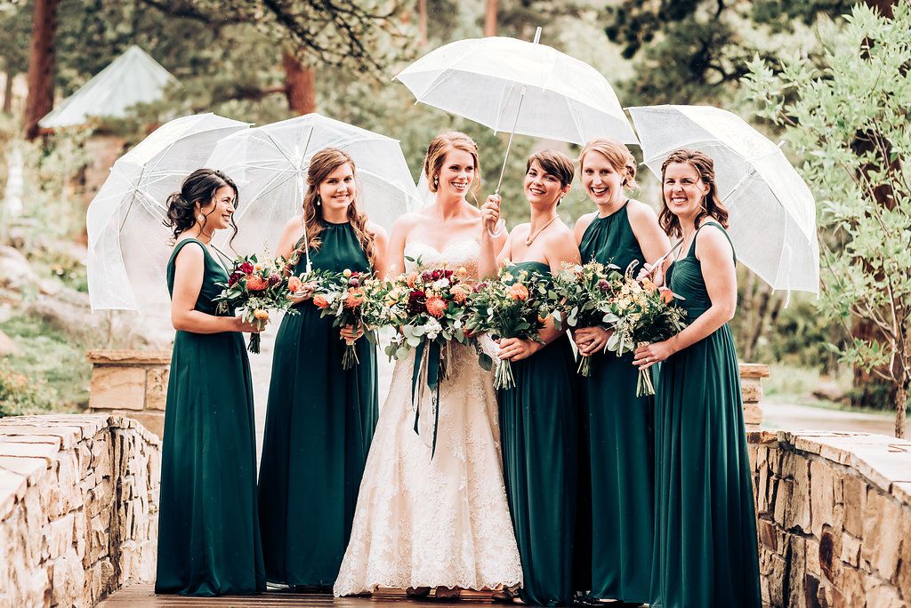 Bridal party in emerald dresses poses with white umbrellas in mountain setting.