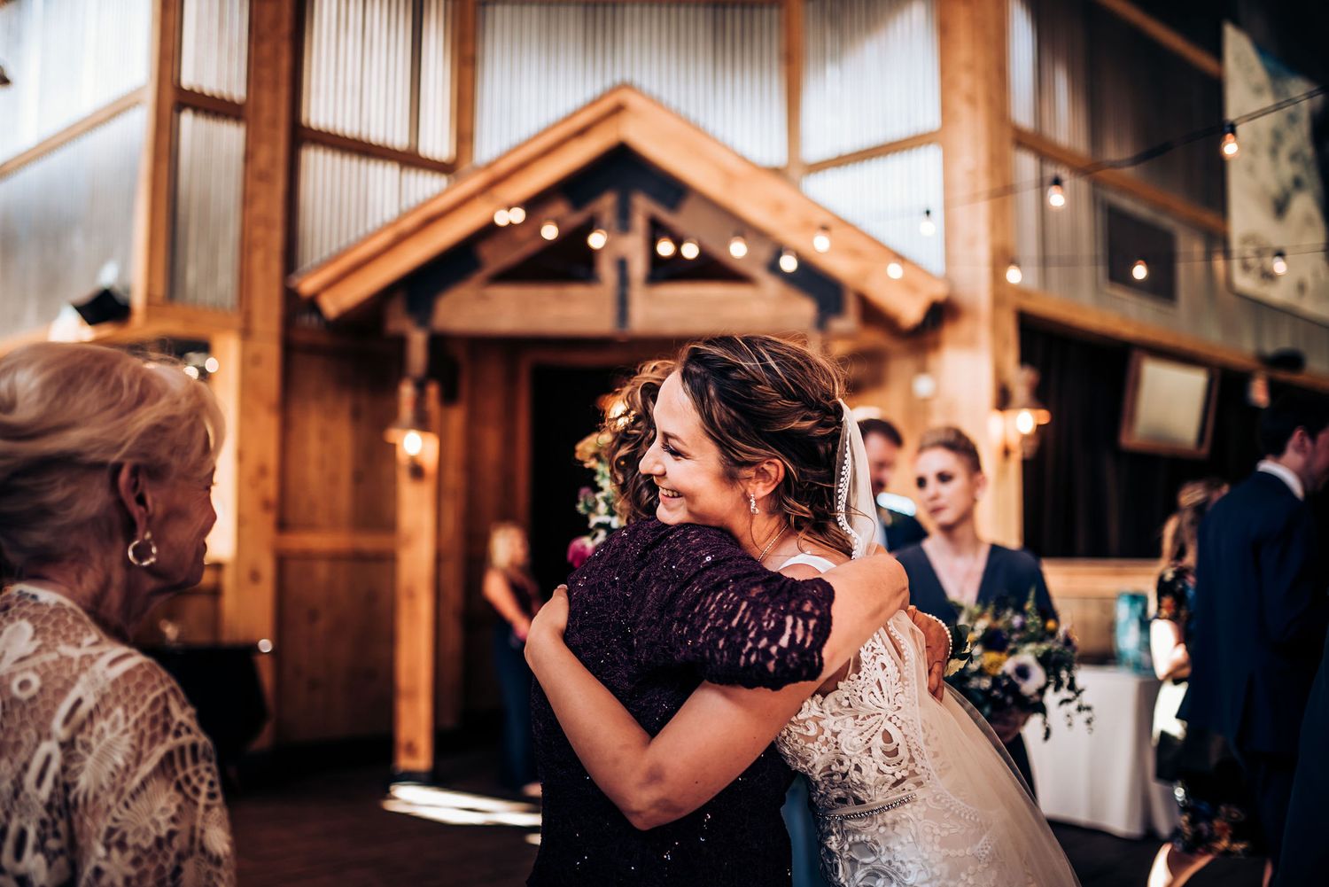 Emotional embrace during wedding reception inside rustic wooden venue with string lights.