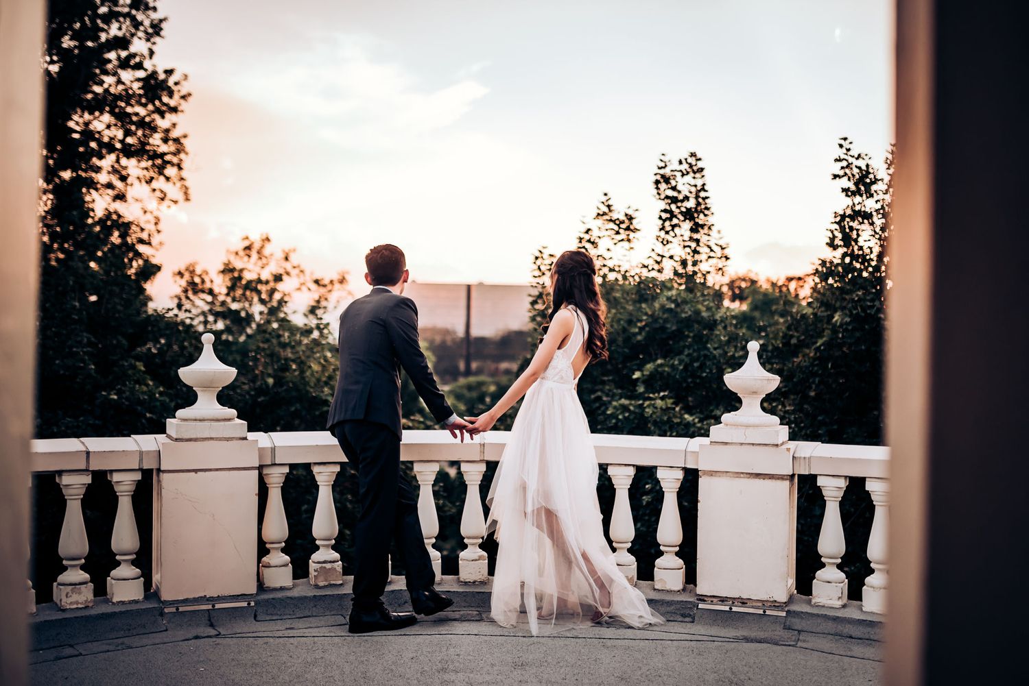 Couple admiring sunset view from elegant white stone balcony during wedding photoshoot.