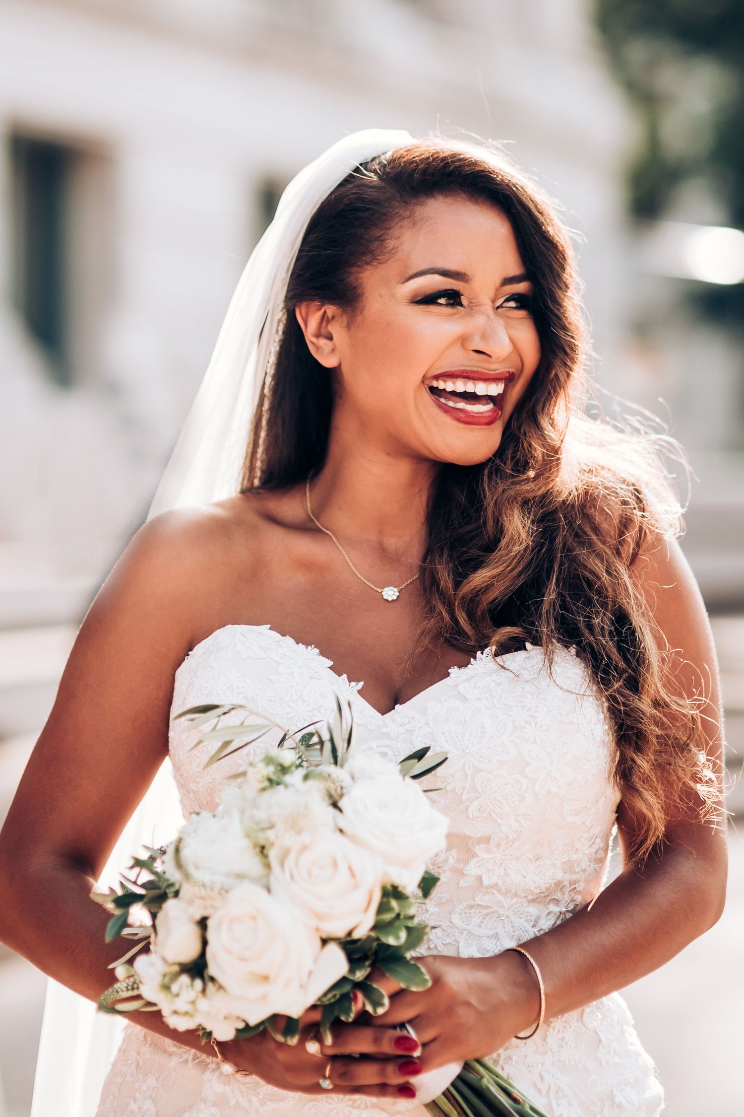 Radiant bride in strapless lace gown holds white rose bouquet and smiles outdoors.