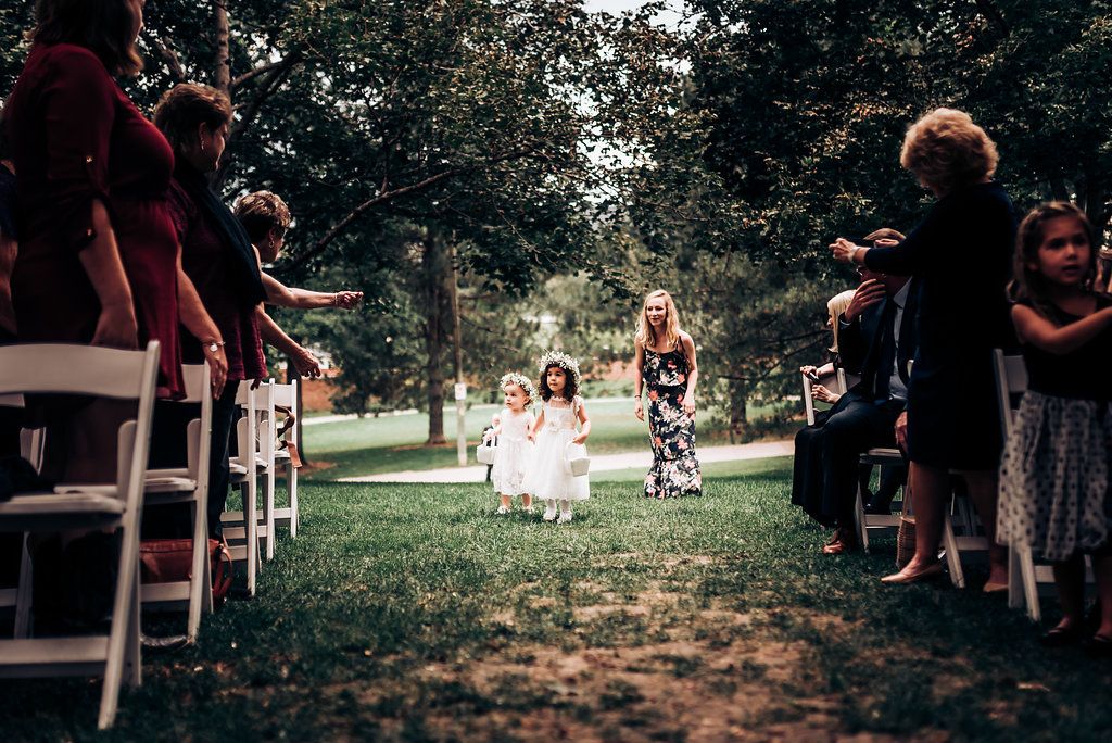 Flower girls in white dresses walk down outdoor wedding aisle between rows of guests.