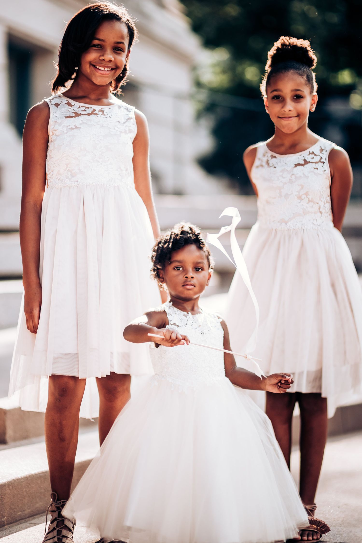 Three young flower girls in white lace dresses pose outdoors for a formal portrait.