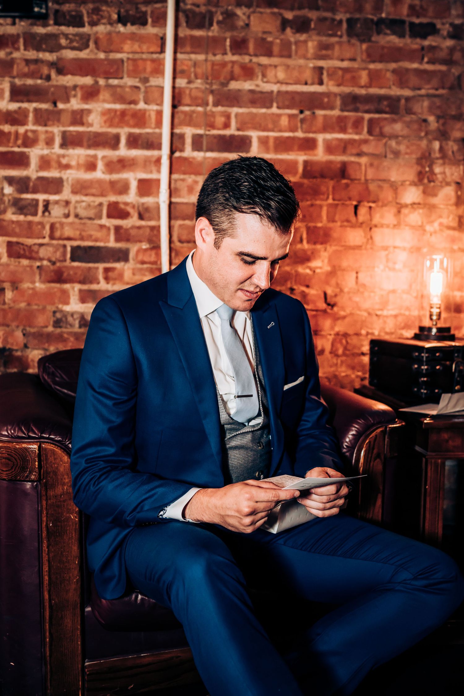 Groom in navy blue suit checks his phone while seated in leather chair against brick wall.