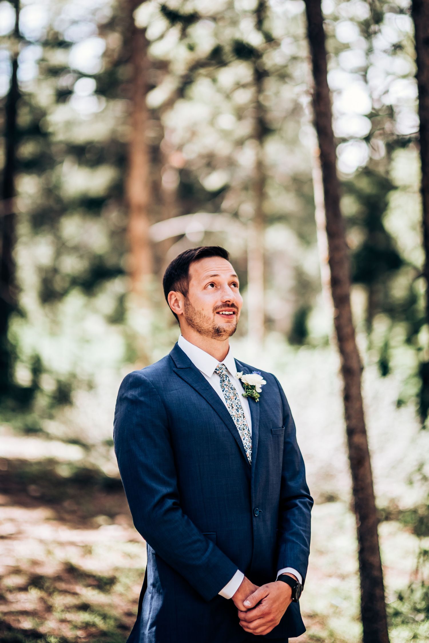 Groom in navy suit and floral tie stands in sunlit forest setting.