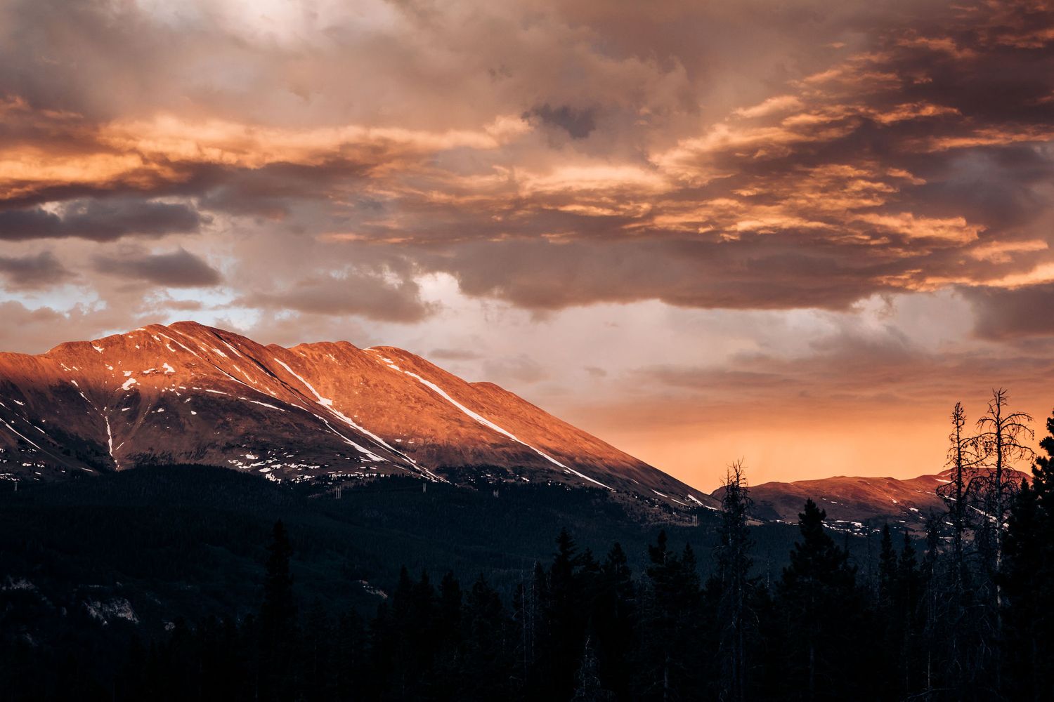 Mountain peak glows orange in dramatic sunset light with stormy clouds overhead.