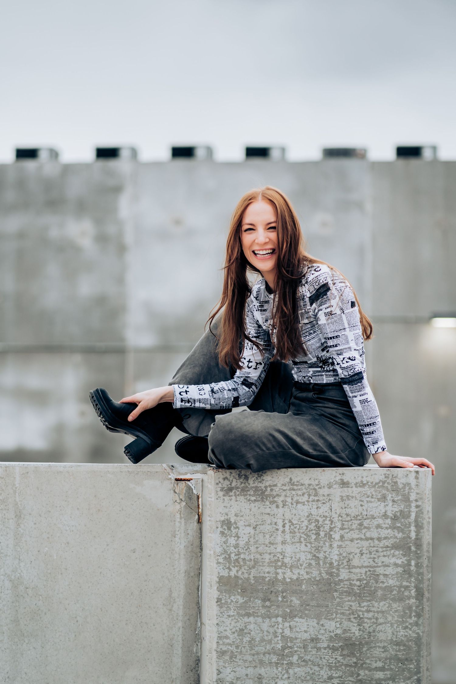 Casual portrait of a woman in newspaper print sweater sitting on concrete wall outdoors.