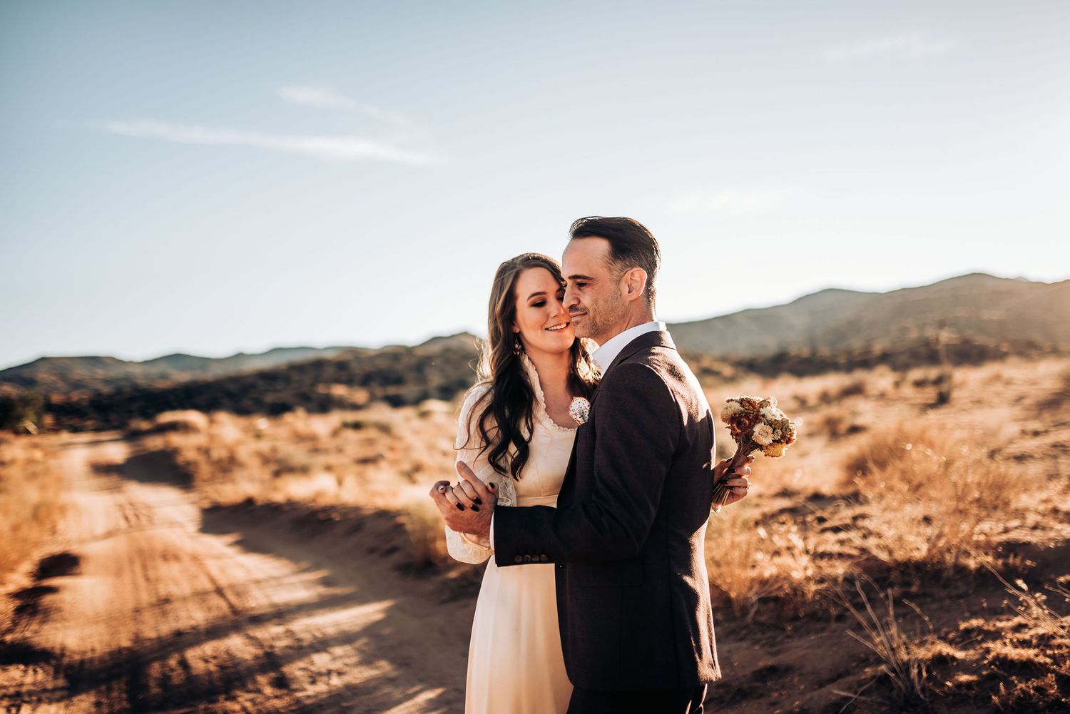 A couple embraces on a desert path with mountains and warm sunlight in the background.