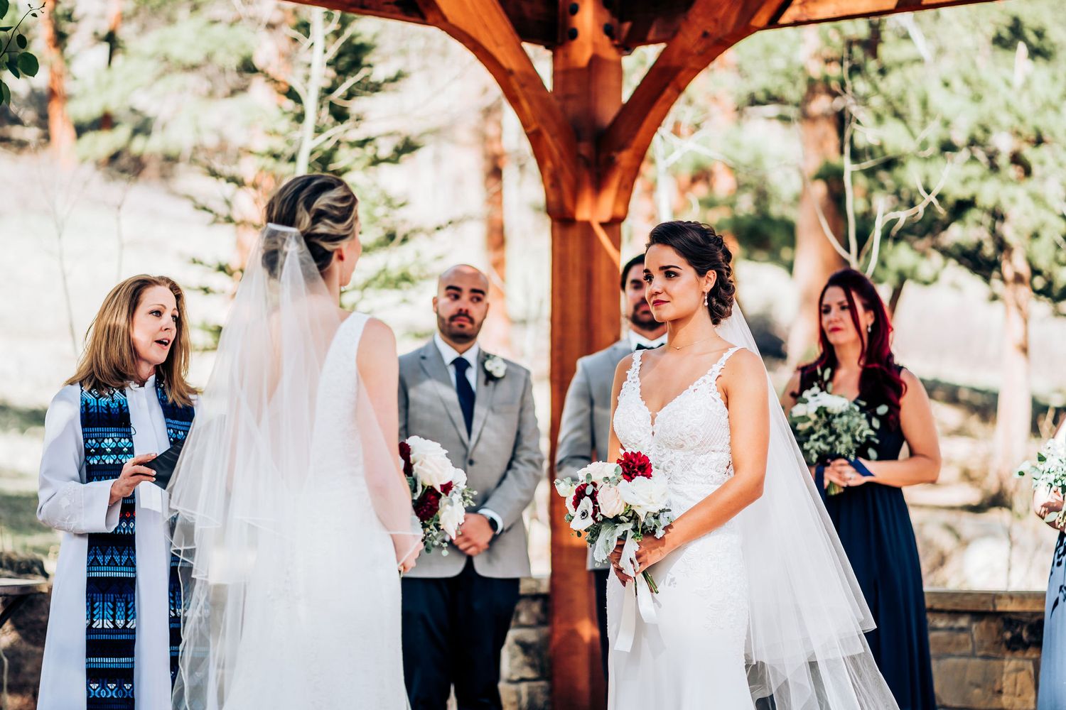 Intimate outdoor wedding ceremony under wooden pergola with wedding party in attendance.