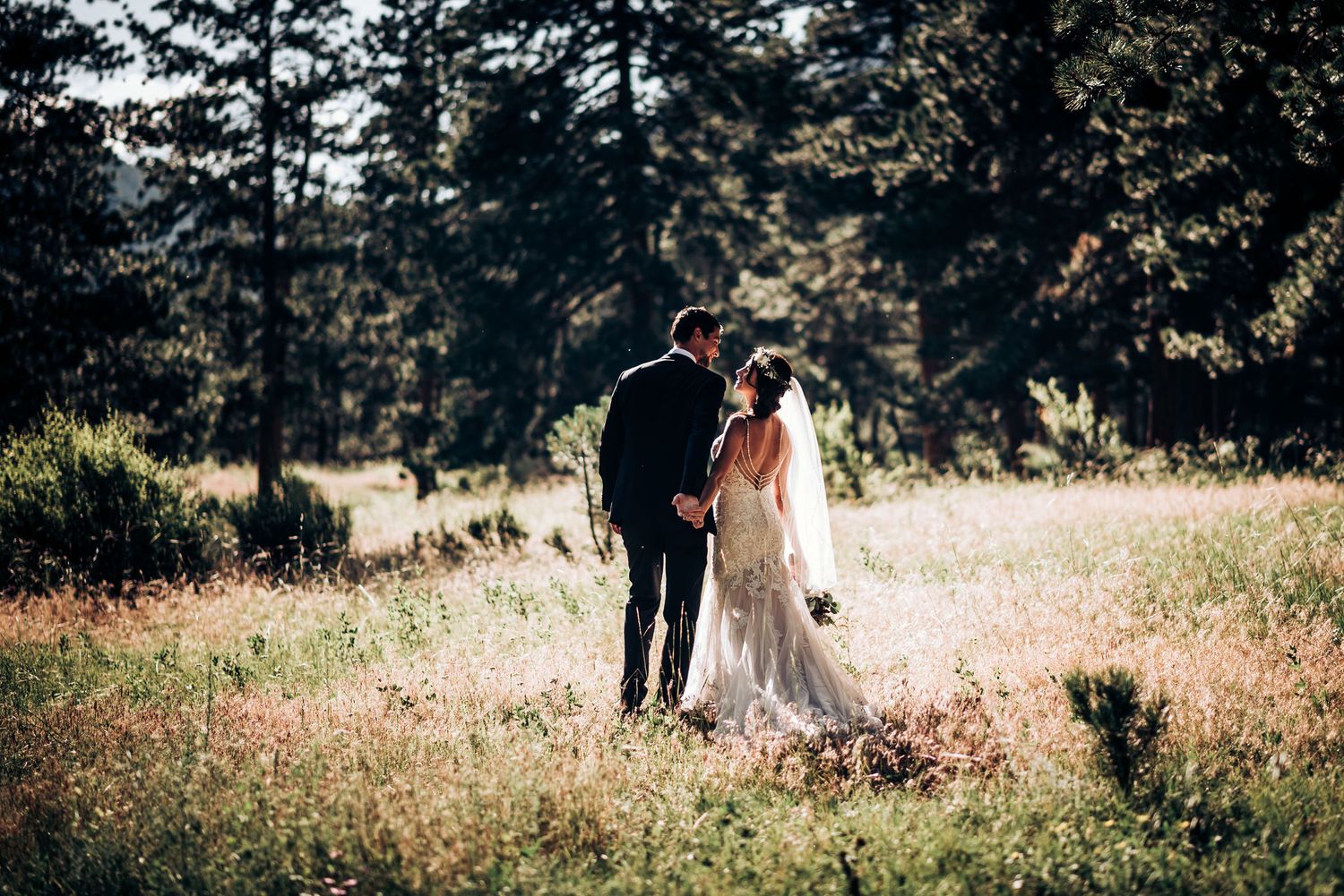 A couple in wedding attire walks through a sunlit mountain meadow.