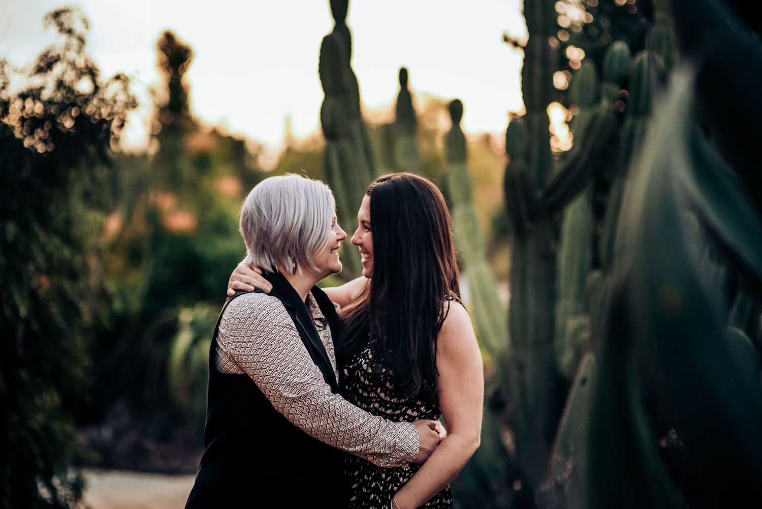 A romantic couple embracing in a desert garden with tall cactus plants during golden hour sunset.