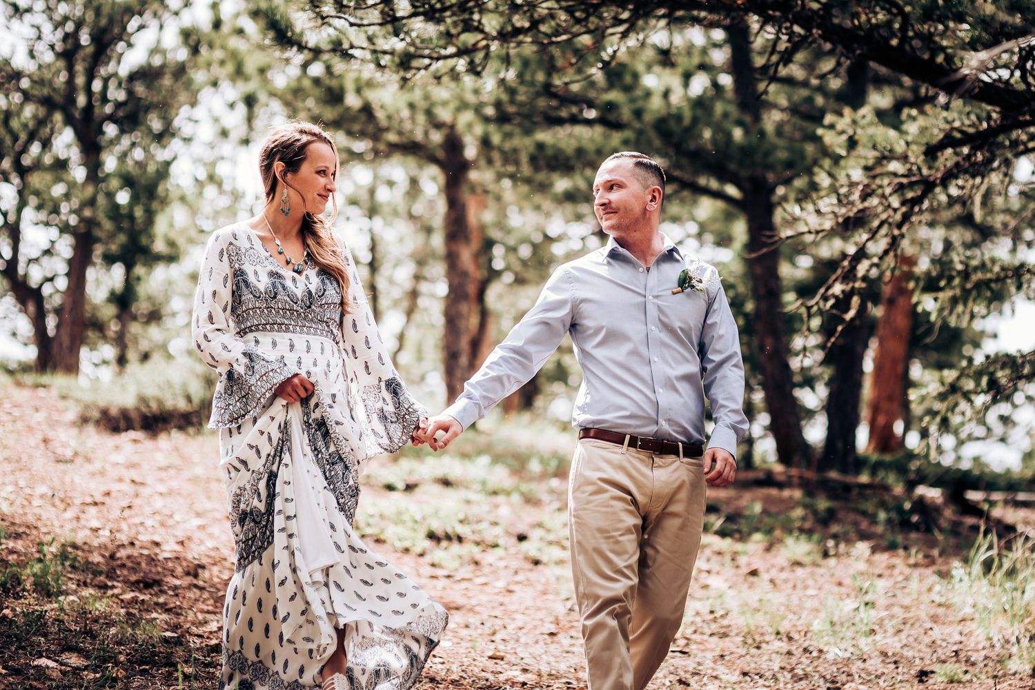 A couple holds hands while walking through a scenic wooded area on a bright day.