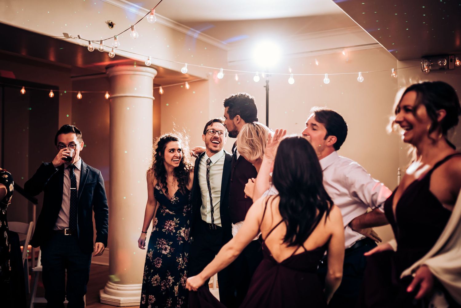 Wedding guests dancing and laughing at a reception under warm string lights.