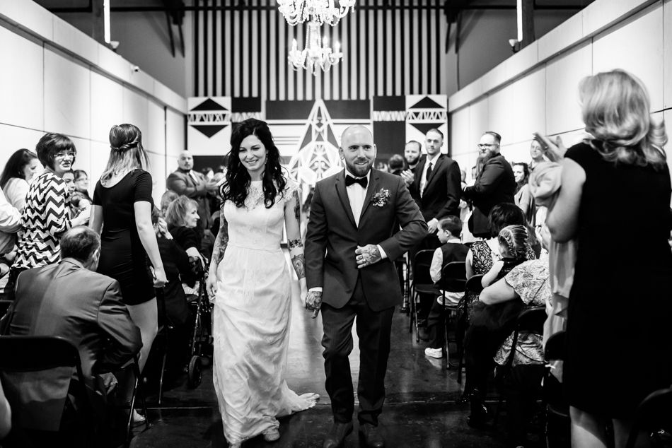 Black and white photo of wedding recessional with guests lining the aisle in an indoor venue.