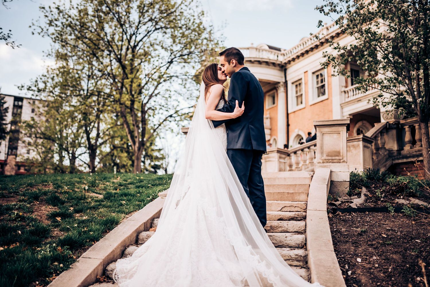 Newlyweds share intimate moment on elegant mansion steps with cathedral veil trailing behind.