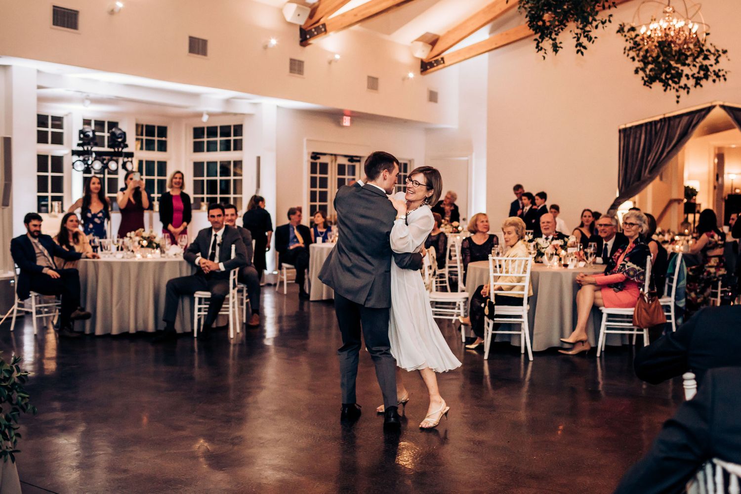 Wedding guests watch a couple dance in an elegant reception venue with round tables.