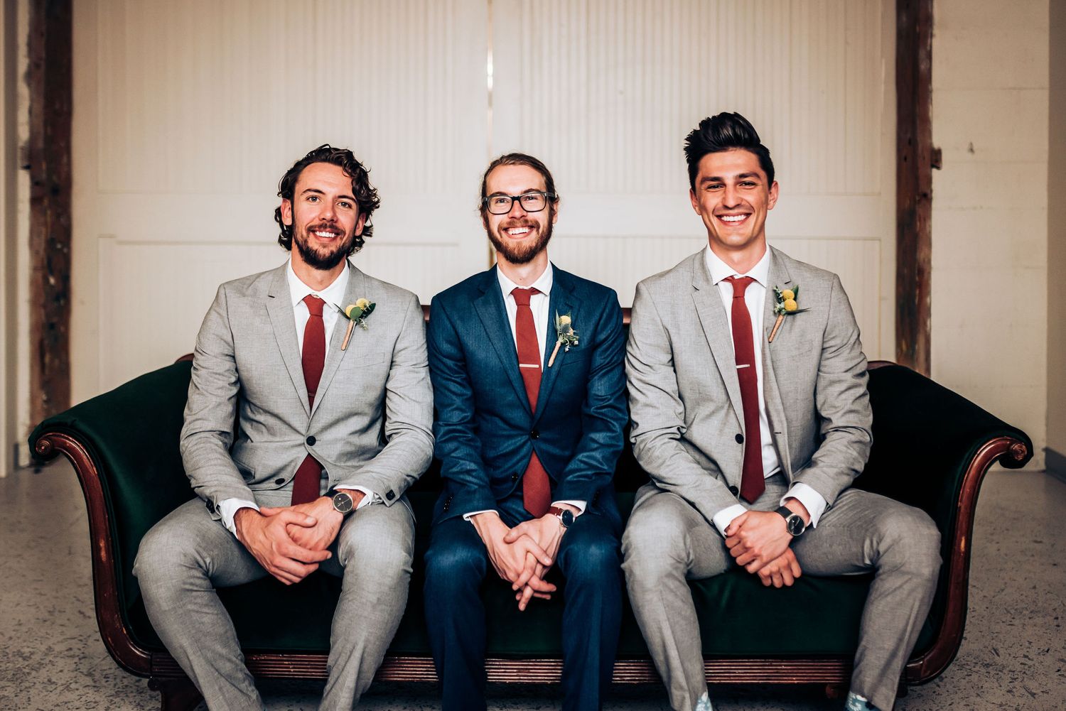 Groomsmen in grey suits and red ties sitting on vintage dark couch.