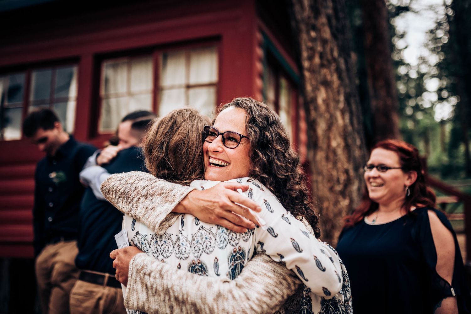 Emotional embrace between friends during an outdoor celebration by a red cabin.