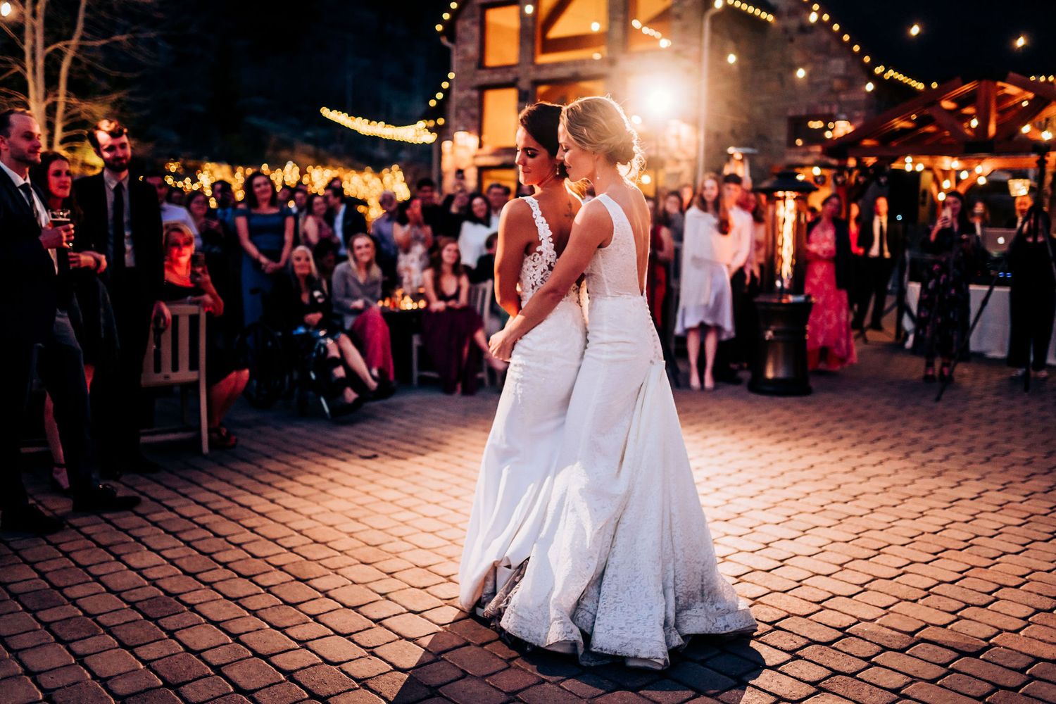 Two people in elegant white dresses share a dance moment at an outdoor evening reception.