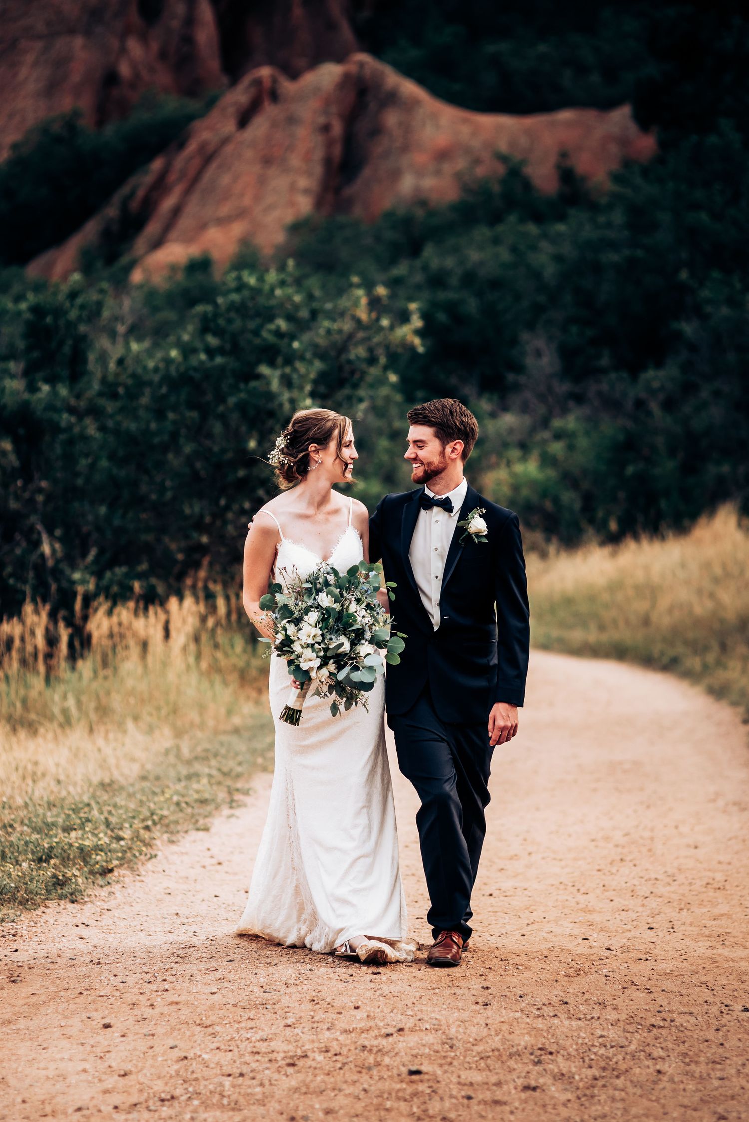 A couple strolls along a winding path with dramatic red rock formations in the background.