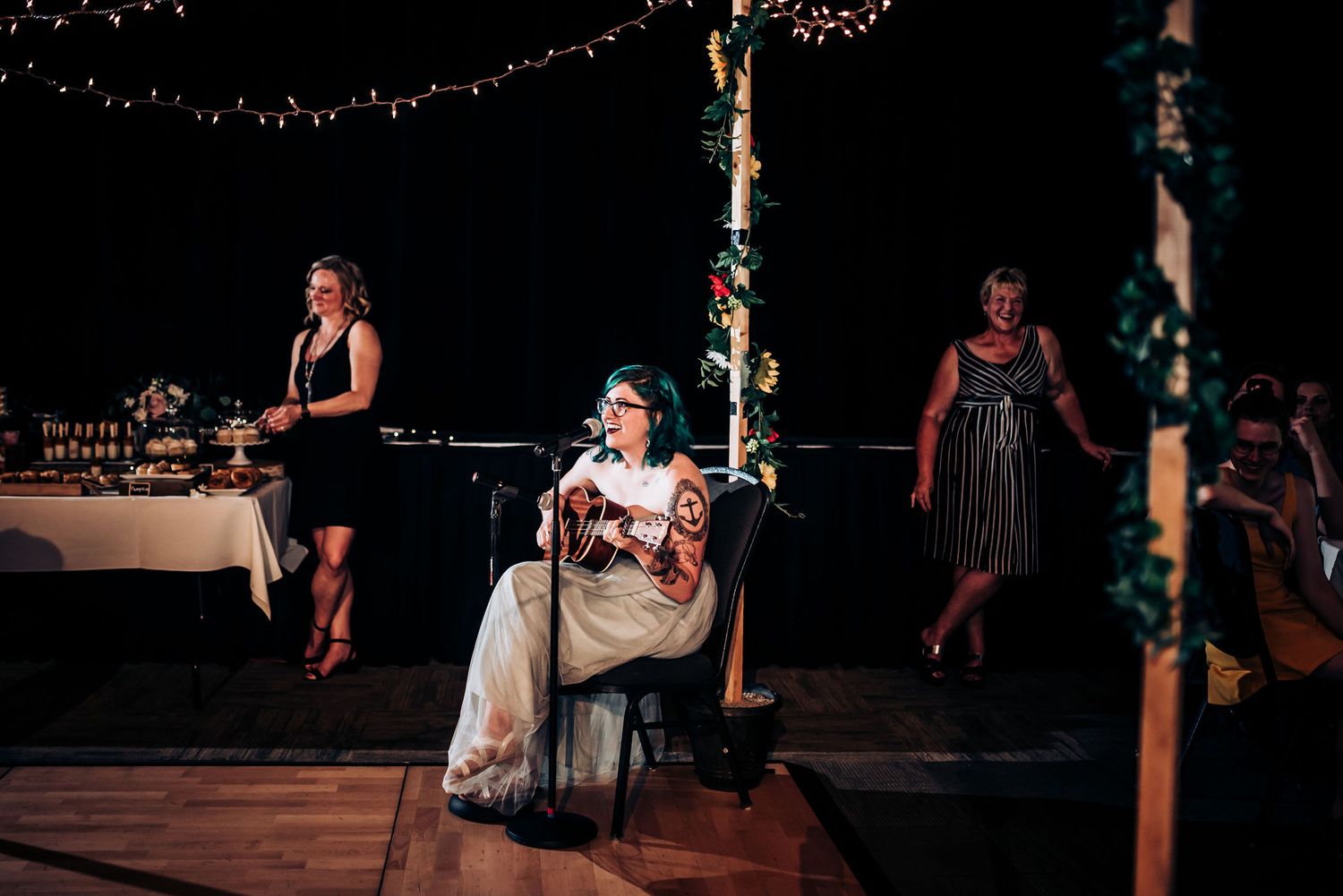 A bride seated at her reception while guests mingle near poles decorated with lights and greenery.