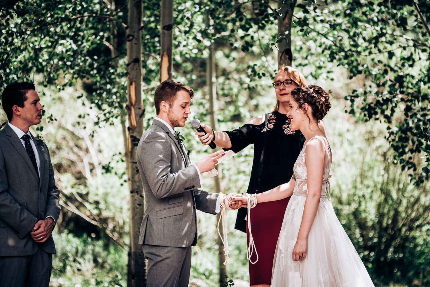 A wedding ceremony takes place in a sunlit forest setting with an officiant presiding over the couple's vows.