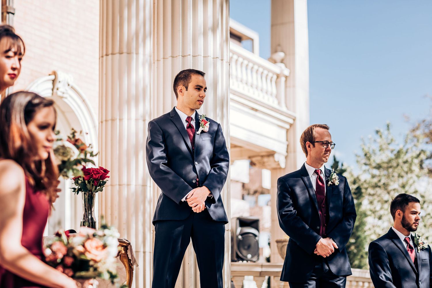 Wedding party members stand in formation during outdoor ceremony with classical columns.