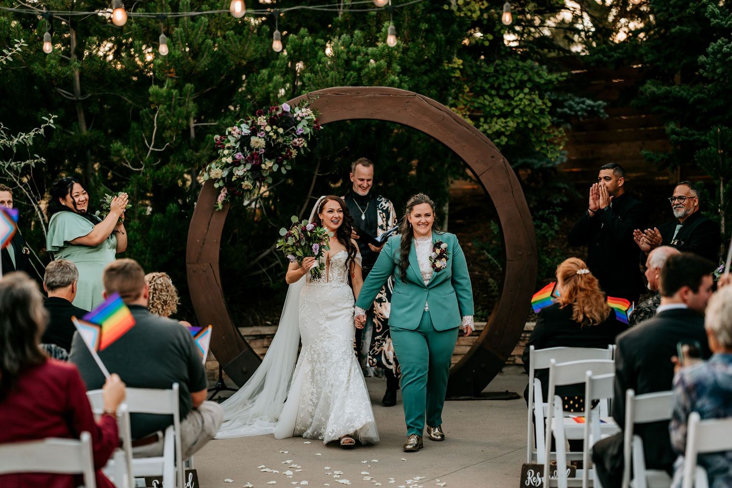 Smiling newlywed couple walks joyfully down the aisle with pride flags waving at their outdoor wedding.