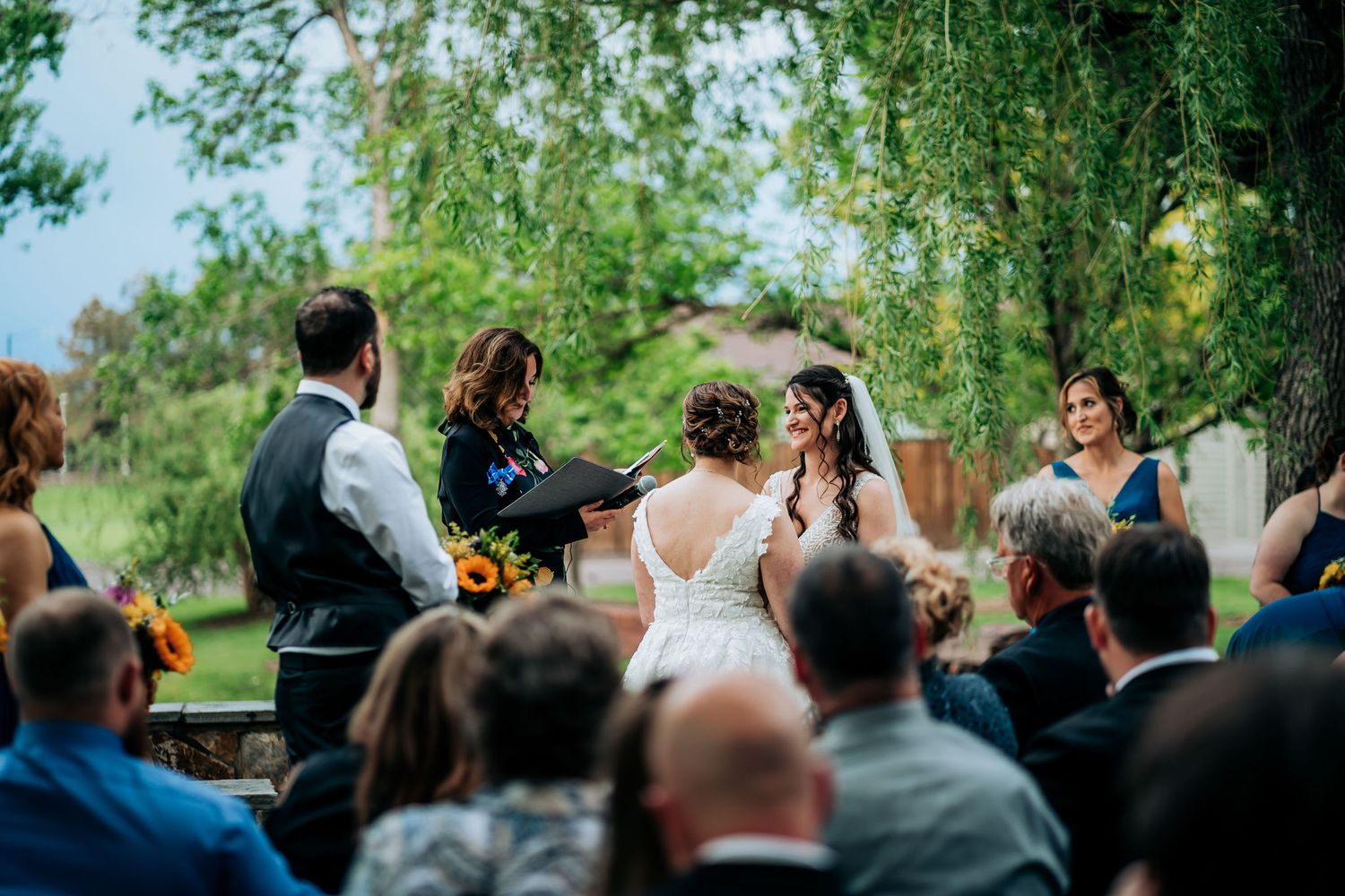 Outdoor wedding ceremony with guests watching couple exchange vows under green trees.