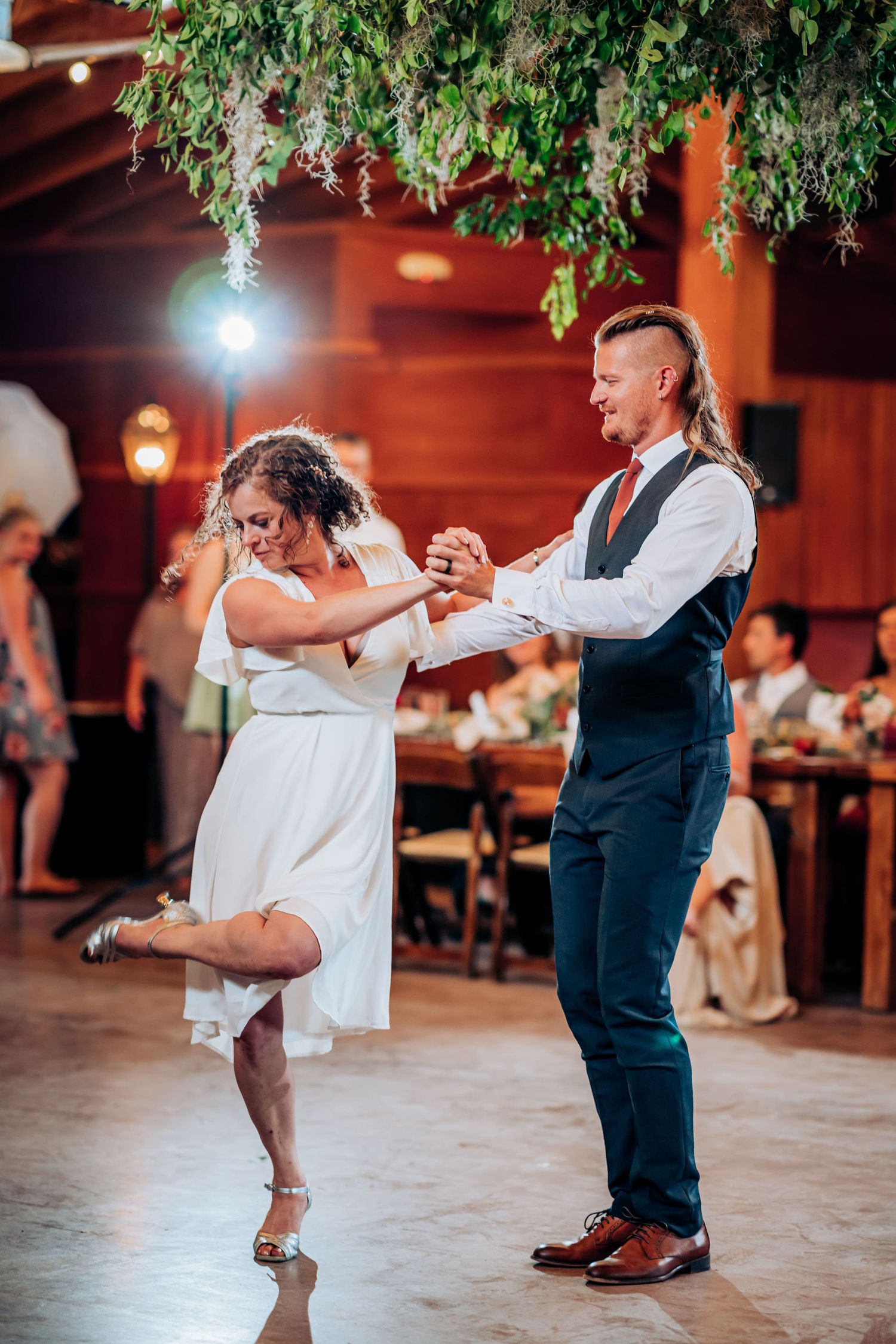 Dancers move energetically on a wooden dance floor under hanging greenery at an indoor venue.
