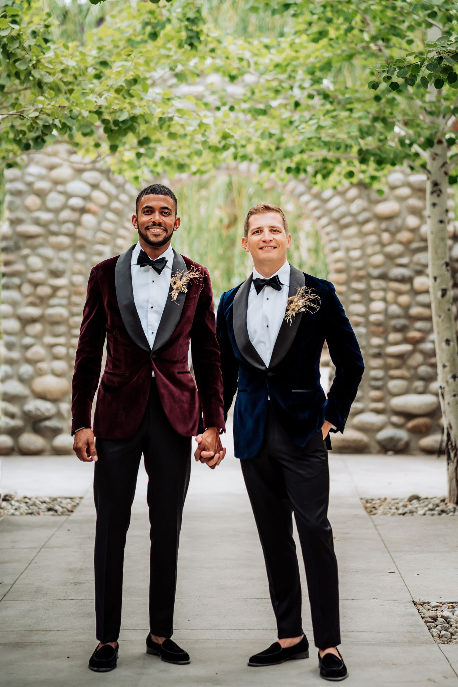 Two people in formal tuxedos hold hands against a stone archway backdrop.