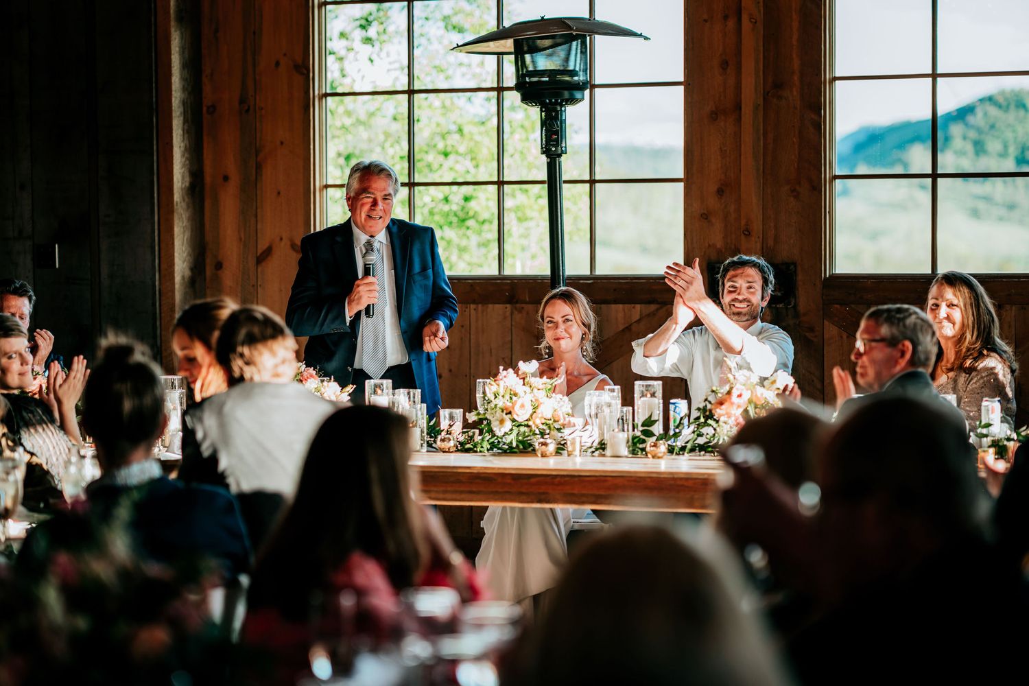 A celebratory dinner speech draws reactions from guests seated at decorated tables in a wooden lodge setting.