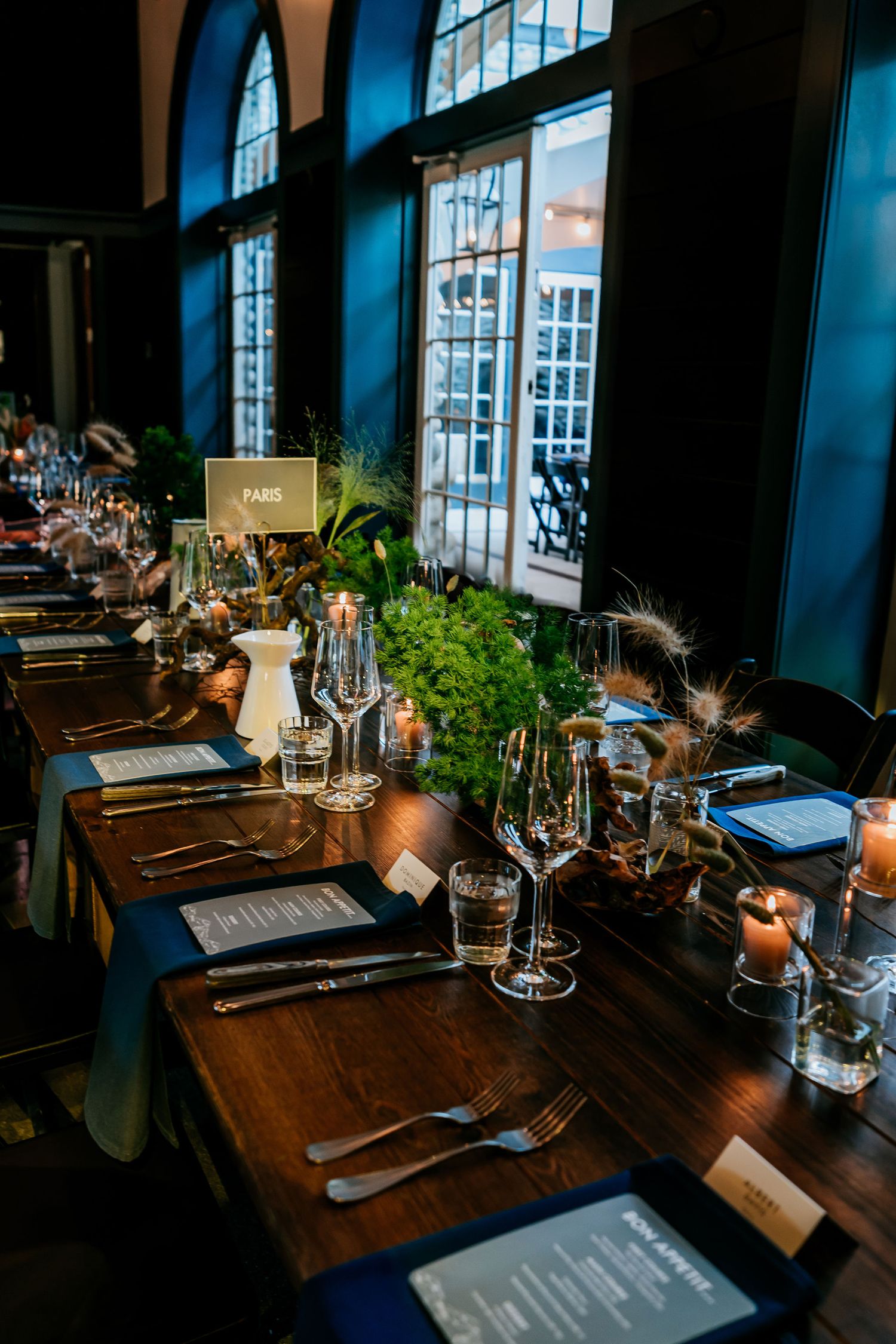 Formal dining table setup with arched windows, greenery, and place settings in moody lighting.