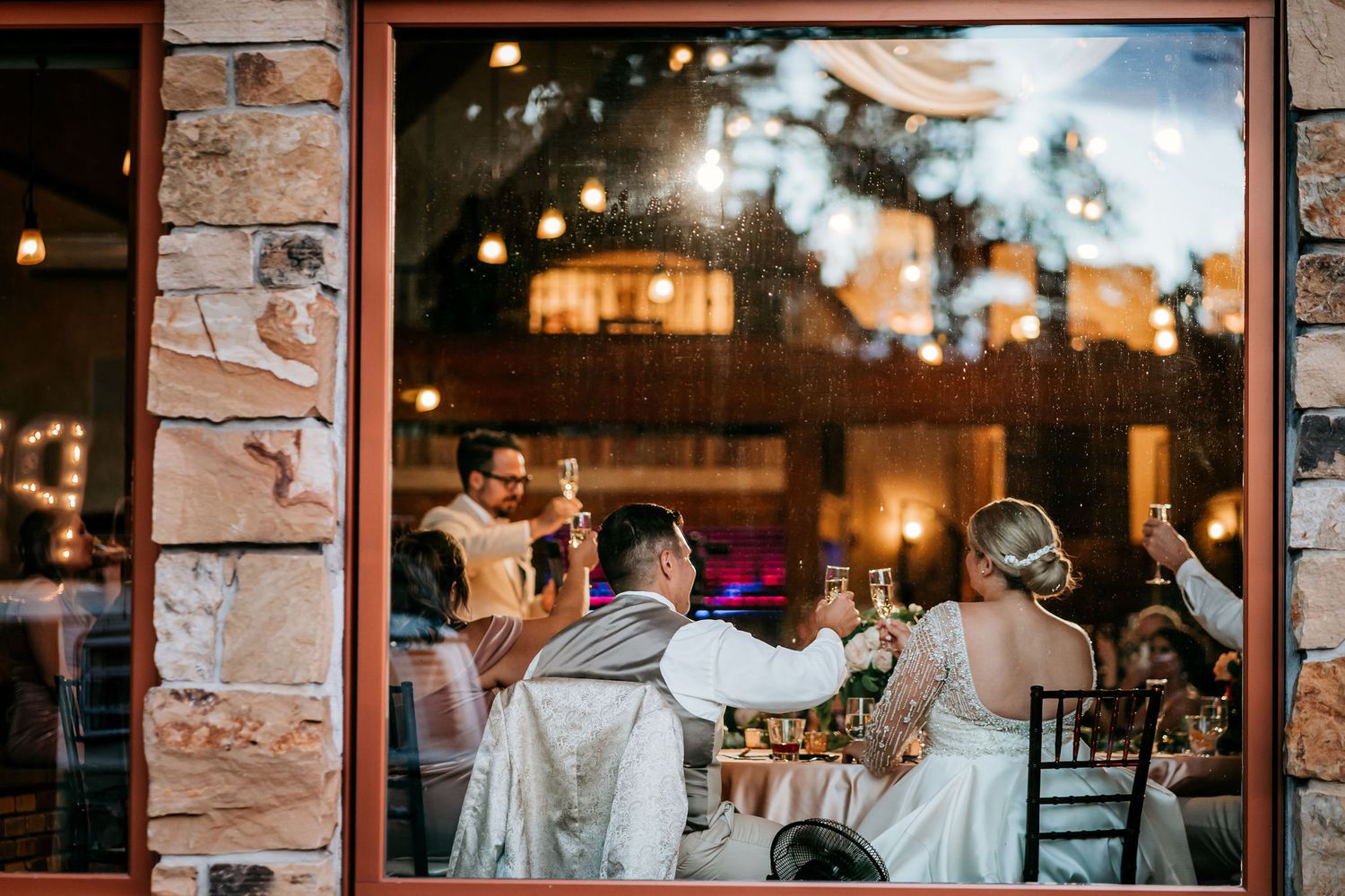 Evening wedding reception viewed through window with stone walls and warm ambient lighting.