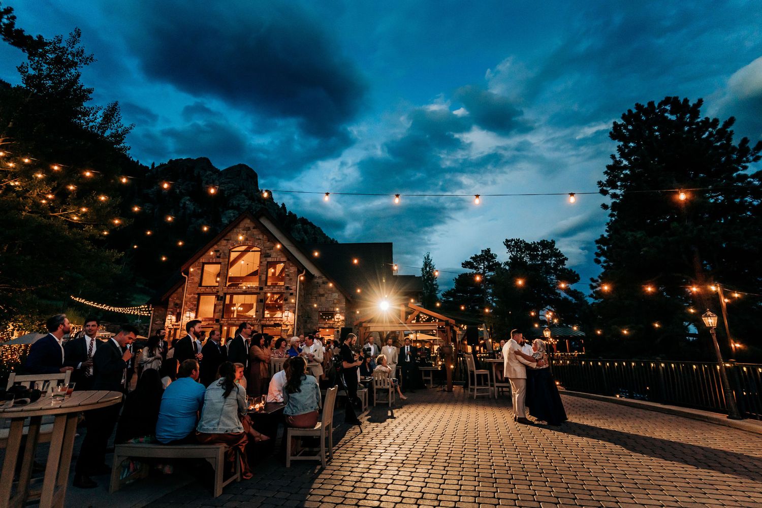 Evening wedding reception at a rustic venue with string lights and dramatic blue sky.