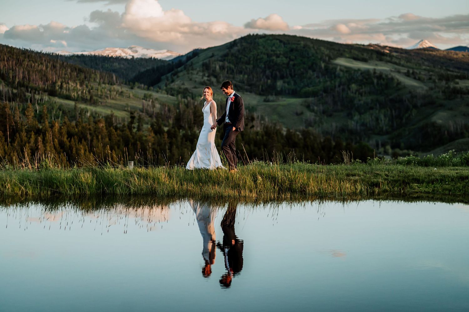 Bride and groom reflected in still mountain lake with snow-capped peaks in background.