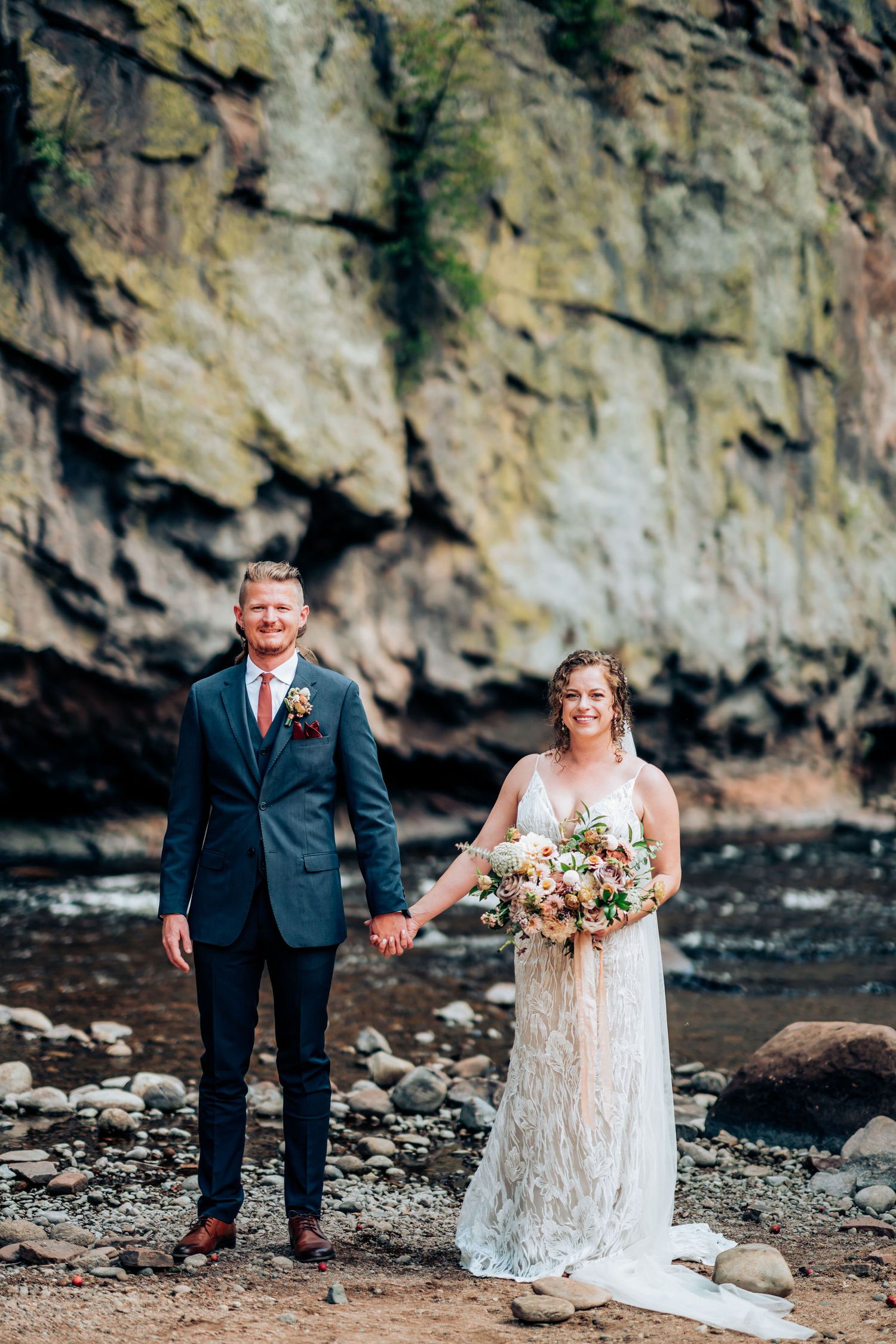 Wedding couple poses in front of towering rock wall by riverside wearing navy suit and lace gown.