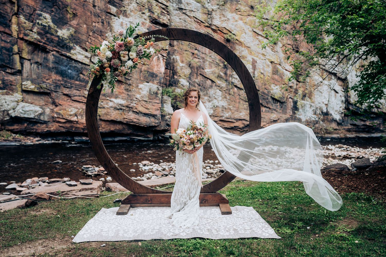 Bride in lace gown poses with flowing veil by circular arch decorated with flowers against rocky backdrop.
