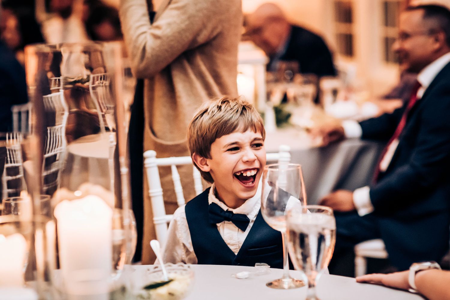 Young boy in bow tie laughs joyfully at wedding reception table.