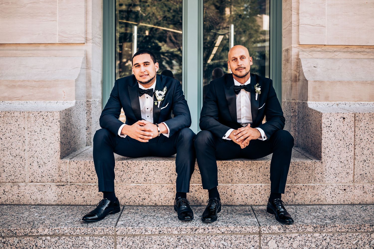 Two people in black tuxedos sit side by side on stone steps at an elegant event.