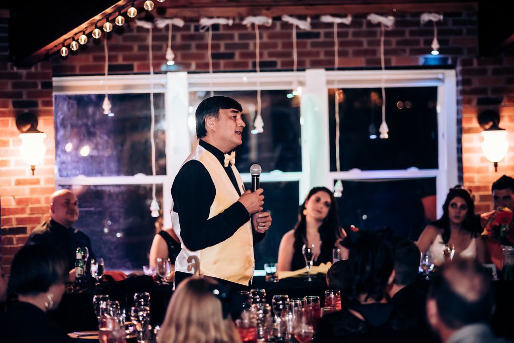 Speaker addressing guests during formal dinner event in brick-walled venue.
