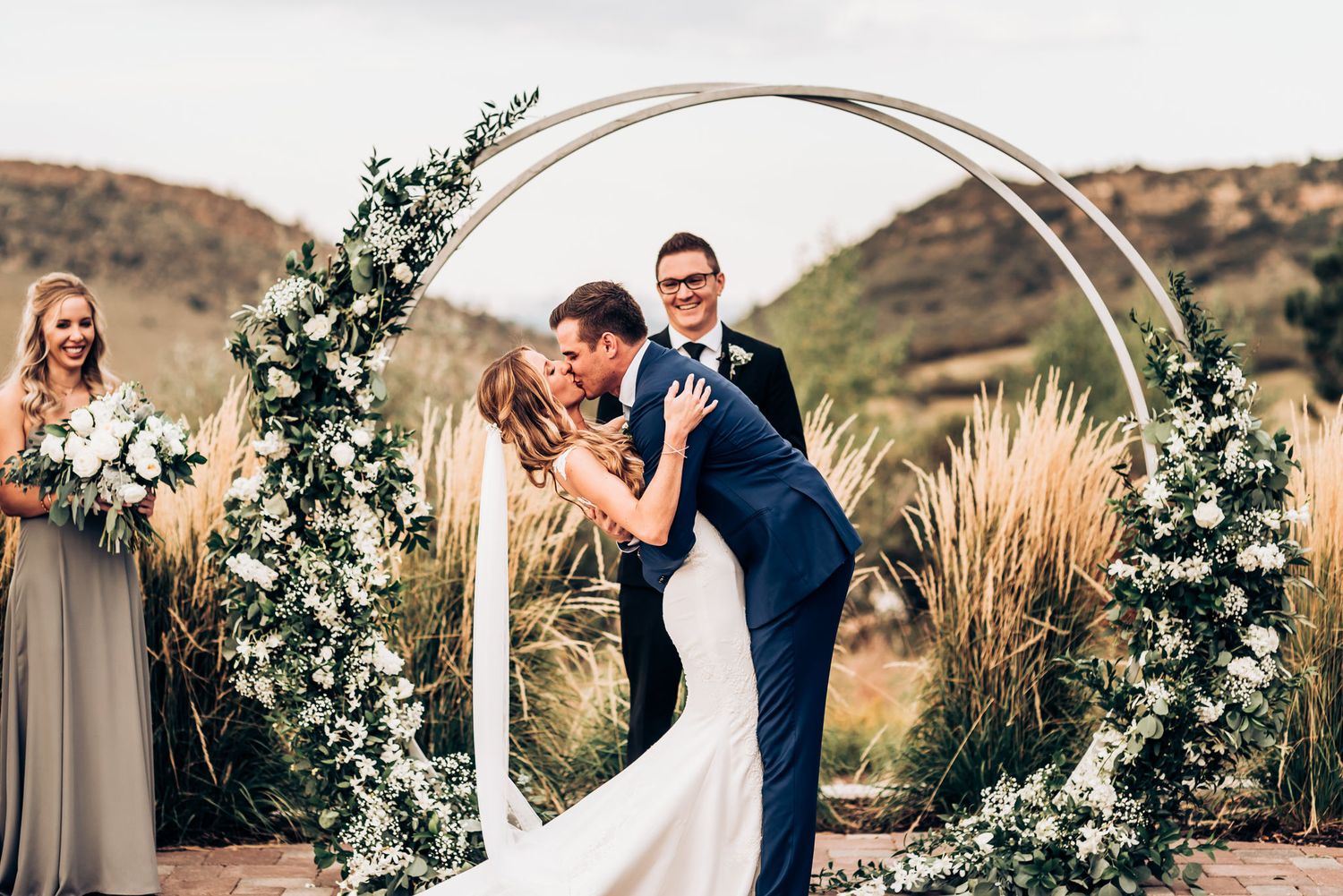 Romantic moment during outdoor wedding ceremony under white flower adorned circular arch.