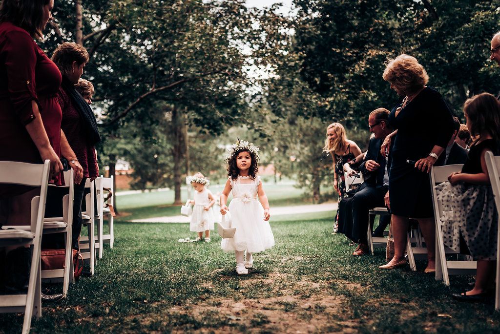 Young flower girl in white dress walks down outdoor wedding aisle between seated guests.